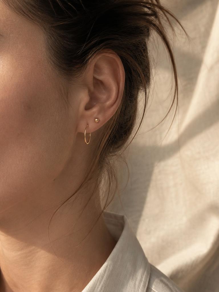 Aretes pequenos de oro en una oreja, luz natural suave sobre fondo beige